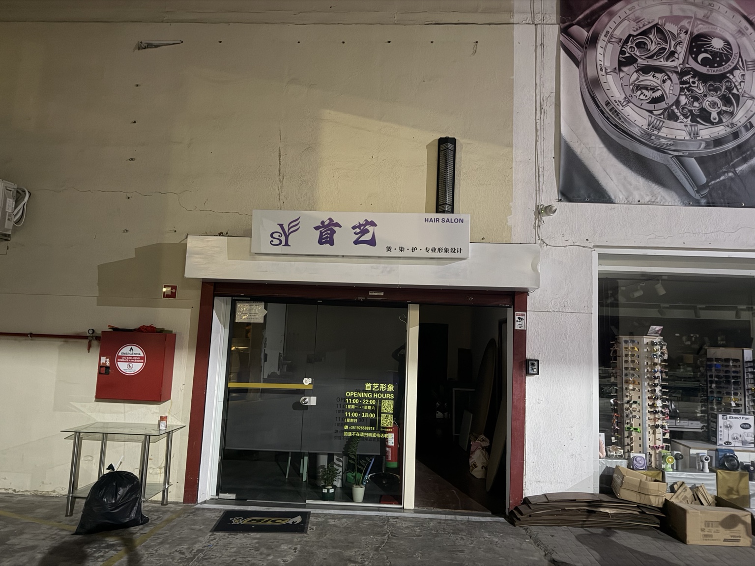 亚洲商城里的首艺理发店,招牌下是玻璃门与半开的入口,旁边摆着纸箱和杂物