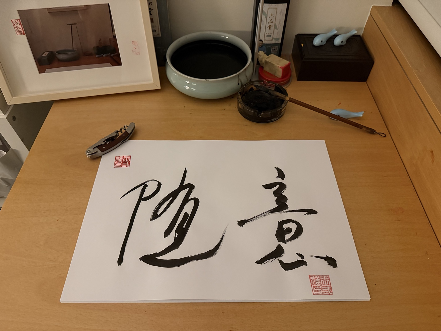 木桌上的毛笔字“随意”,纸上有红色印章,旁边放着毛笔、笔洗与开瓶器