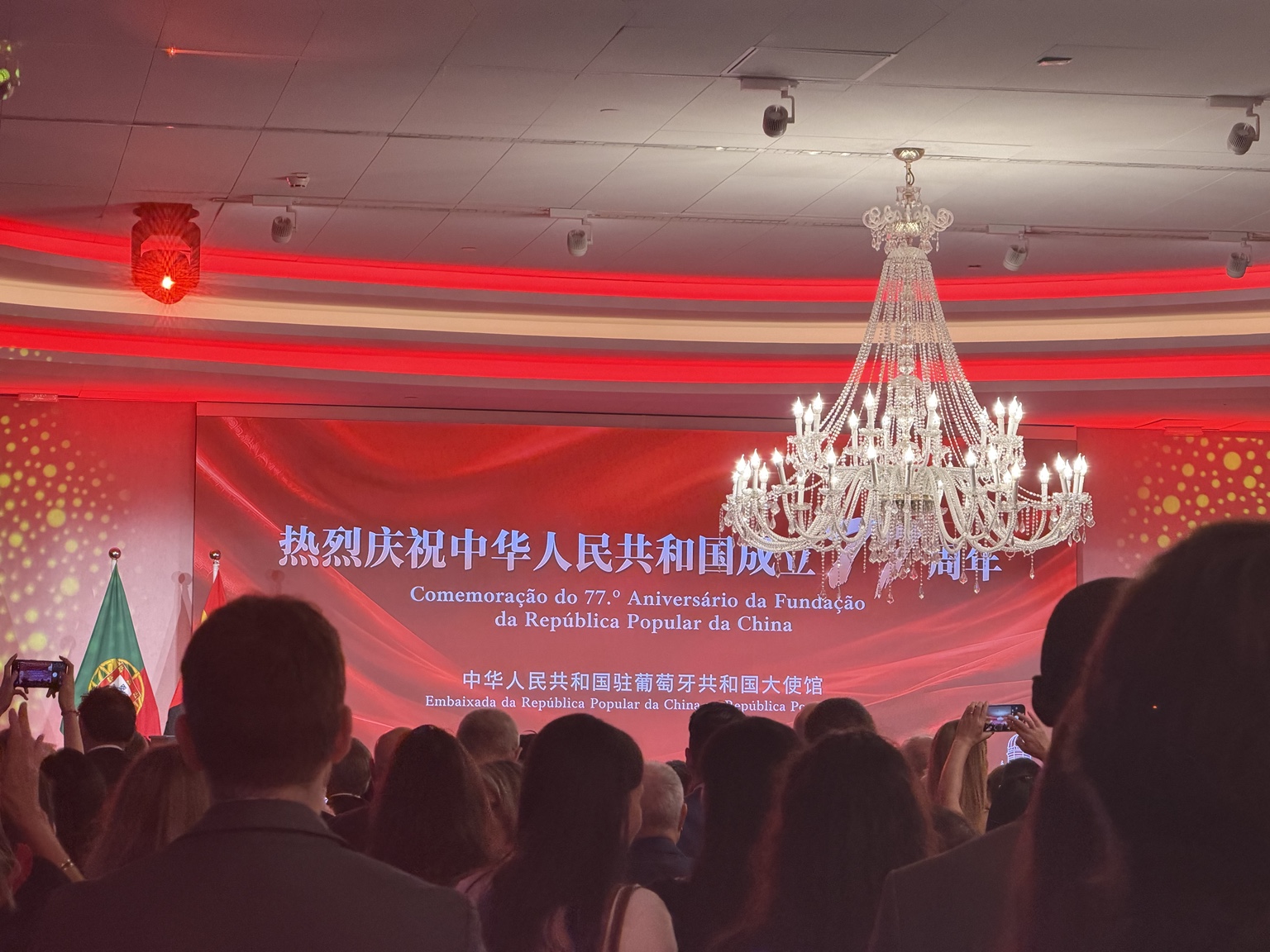 国庆活动现场,红色背景板上写着庆祝中华人民共和国成立77周年,中葡国旗与水晶吊灯映衬宾客