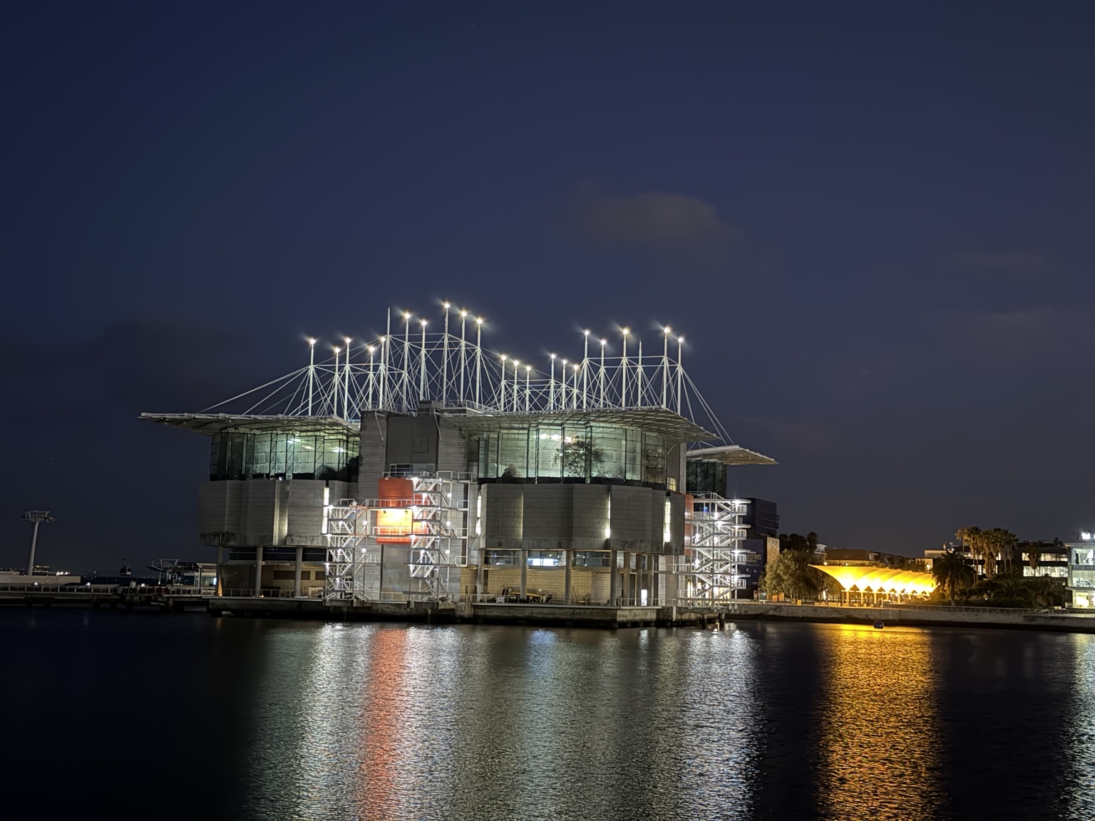 夜幕下水族馆的立杆与拉索亮起白灯，建筑和右侧遮阳伞的暖色灯光倒映在水面上