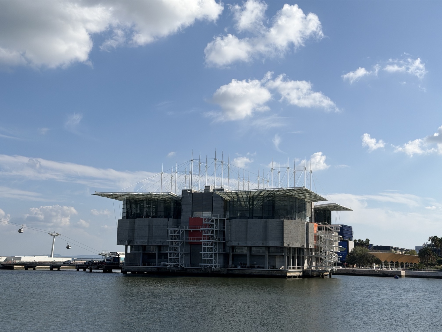 水面上的里斯本水族馆，灰色墙壁与玻璃立面，上方是伸展的屋顶、立杆和拉索，左侧远处有两辆缆车